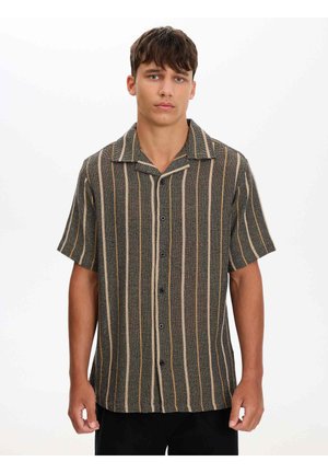 Joven con camisa negra de manga corta con rayas verticales beige y naranja, de pie frente a un fondo blanco liso.