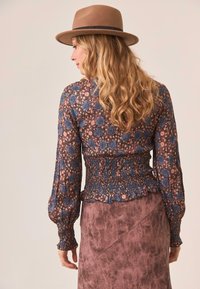 Blusa floral en tonos de azul y rosa, con cintura fruncida y mangas largas abullonadas, combinada con una falda texturizada rosa y un sombrero marrón.