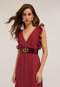 Rotes fließendes Kleid mit Rüschensäumen, in der Taille von einem glänzenden kastanienbraunen Gürtel mit einer goldenen ovalen Schnalle gerafft. Glatte Stoffoberfläche.
