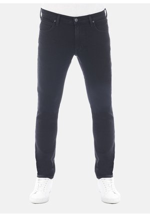 Mężczyzna w czarnych dopasowanych dżinsach i białych trampkach, stojący na białym tle.