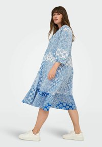 Modra obleka s potiskom in tremi četrtinastimi rokavi, ki vključuje zapletene vzorce v svetlejši in temnejši modri barvi. Kombinirano z belimi supergami.