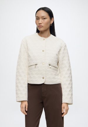 Femme aux cheveux noirs et raides portant une veste matelassée blanche avec des fermetures éclair devant et un pantalon marron sur un fond uni clair.