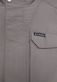 Veste Columbia grise avec une texture cousue, des poches latérales et une fermeture à pression. Logo de la marque affiché sur la poche avant.