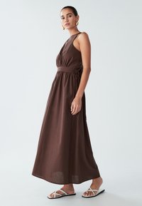 Sieviete, tērpusies bezpiedurkņu brūnā maksi kleitā ar sazvērestības jostasvietu un baltām siksnām plakanām sandalēm, stāv pretī vienkāršai gaišai fona.
