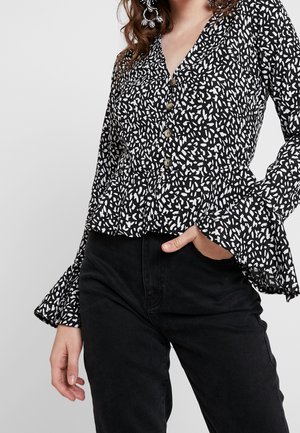 Schwarze Bluse mit Knopfleiste, die ein weißes abstraktes Blattmuster, strukturierte Stoffe, einen Peplum-Saum und ausgestellte Ärmel aufweist, kombiniert mit schwarzen Hosen.