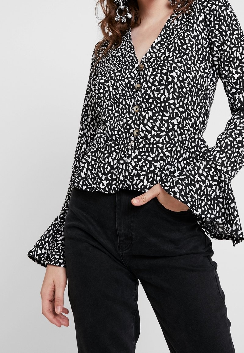 Blusa nera con bottoni a maniche lunghe, caratterizzata da un motivo astratto a foglie bianche, tessuto textured, orlo a peplo e maniche svasate, abbinata a pantaloni neri.