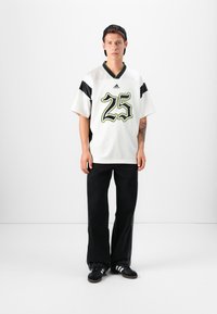 White short-sleeve jersey with black accents and a green-outline '25' logo, paired with black wide-leg pants and black sneakers.