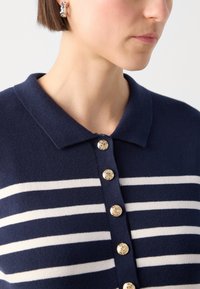 Námořnický modrý pruhovaný cardigan s bílými vodorovnými pruhy, s límcem a šesti dekorativními zlatými knoflíky vpředu.