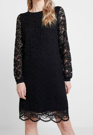 Vestido negro de encaje hasta la rodilla con mangas largas transparentes, bajo festoneado y escote redondo en un modelo de pie.