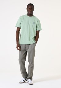 Lichtgroen katoenen T-shirt met een ronde hals en logo op de borst, gecombineerd met grijze broeken en witte sneakers met zwarte strepen.