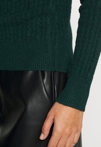 Suéter de canalé verde oscuro con mangas largas, hecho de una textura suave, combinado con pantalones de cuero negros. Acercamiento de una mano sobre el dobladillo del suéter.