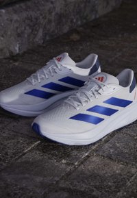 Chaussures de sport blanches avec des rayures bleues, dessus texturé, semelle matelassée et détail rouge au niveau du talon. Convient pour le sport et les tenues décontractées.