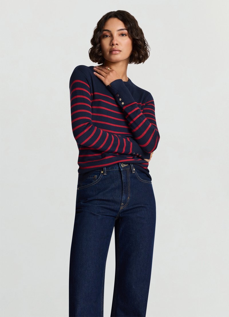 Navy striped sweater with red stripes, round neck, long sleeves, and button accents at the cuffs, paired with dark blue wide-leg jeans.