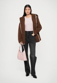 Frau trägt eine braune Fleecejacke, ein hellrosa Oberteil, schwarze Jeans und schwarze Schuhe, während sie eine hellrosa Handtasche vor einem schlichten Hintergrund hält.
