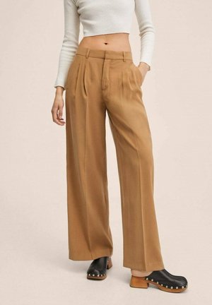 Kobieta ubrana w camelowe, szerokie spodnie z wysokim stanem i plisami, białą, krótką bluzkę z długim rękawem oraz czarne chodaki na drewnianym obcasie.