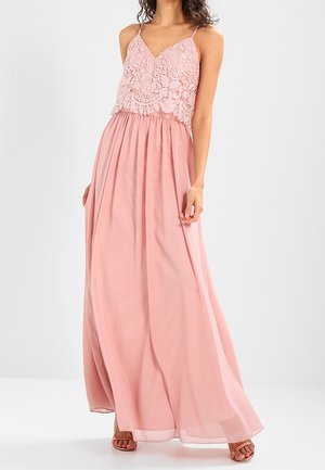 Vrouw in een mouwloze roze maxi-jurk met kanten lijfje en vloeiende rok, gecombineerd met sandalen met open teen.