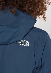 Personne aux cheveux bouclés portant une veste à capuche bleue avec le logo blanc The North Face dans le dos près de l'épaule.