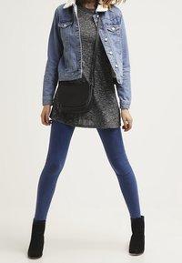 Giacca di jeans con colletto in pelliccia, tunica grigia, leggings blu e stivaletti neri alla caviglia. L'outfit è completato da una borsa a tracolla nera.