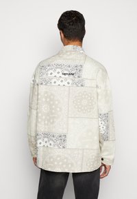 Camicia a maniche lunghe con design a patchwork in beige chiaro e nero, caratterizzata da motivi paisley e un logo sulla parte superiore della schiena.