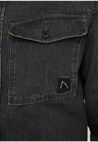 Giacca di jeans nera con taschino sul petto con bottone. Il tessuto ha una texture sottile e include una toppa nera con logo sulla tasca.