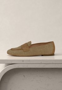 Ανοιχτό καφέ υφασμάτινο μοκασίνι με σουέντ λεπτομέρειες και επίπεδη σόλα, τοποθετημένο σε επιφάνεια από ανοιχτόχρωμη πέτρα, με ουδέτερο φόντο.