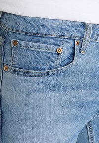 Jeans de mezclilla azul claro con un bolsillo delantero con herrajes dorados y costuras amarillas en contraste. La textura es suave con un acabado desgastado.