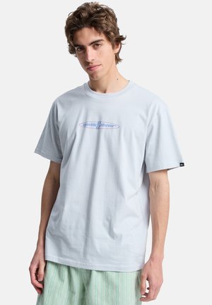 Jasnoniebieska bawełniana koszulka z krótkim rękawem, z centralnym logo Quiksilver w kolorze fioletowym, noszona z wzorzystymi, miętowo-zielonymi szortami.