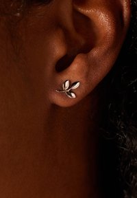 Zilveren bladvormige studoorbel met drie bloemblaadjes-achtige elementen, tegen een gestructureerde huidachtergrond. Close-up weergave benadrukt de details.