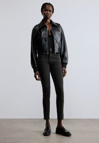 Chaqueta corta de cuero negro con acabado liso, combinada con jeans ajustados oscuros y mocasines negros, exhibida sobre un fondo neutro.