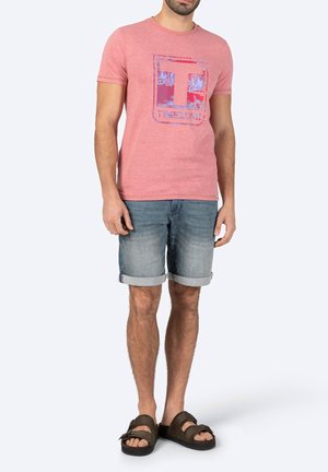 Rotes T-Shirt mit kurzen Ärmeln und einem grafischen Logo in Blau und Lila, kombiniert mit hellblauen Denim-Shorts, die am Saum umgeschlagen sind, und braunen Sandalen.