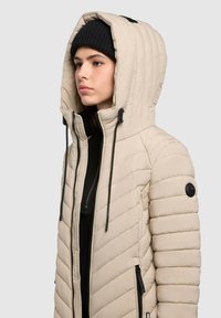 Beige isolierte Jacke mit Kapuze, gestepptem Design, schwarzem Reißverschluss und Kordelzügen, mit seitlichen Taschen und einem runden Logo-Patch.