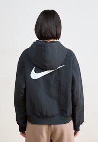 Melnas kapuces jaka ar lielu baltu Nike logo aizmugurē, ar brīvu piegriezumu un ribotām piedurknēm un apakšmalu.