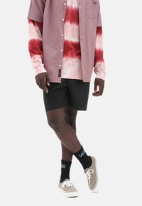 Camisa de manga larga de estilo tie-dye rojo y rosa debajo de una camisa de botones gris de manga corta, pantalones cortos negros y zapatillas color beige con suelas blancas y calcetines negros.