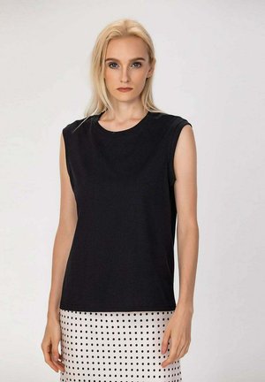 Femme blonde portant un haut noir sans manches et une jupe blanche à pois noirs, debout devant un fond clair uni.