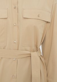 Beige button-up shirt with two breast pockets, belted waist, and smooth fabric texture. Features large buttons and a relaxed fit design.