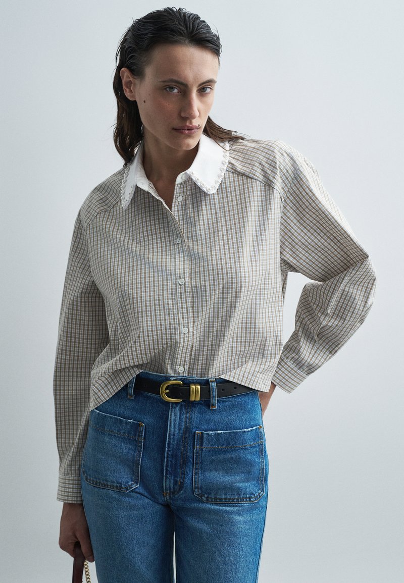 Beige and brown plaid button-up shirt with a white collar, paired with high-waisted blue jeans and a black belt with a gold buckle.