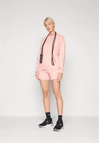 Camisola com capuz rosa claro com cordões, calções combinando, ténis pretos e meias. A camisola com capuz tem um bolso frontal e uma tira ajustável.