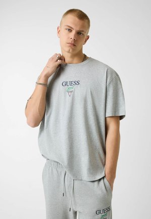 Giovane uomo che indossa una maglietta grigio chiaro del marchio Guess e pantaloni della tuta coordinati, posando con una mano vicino al collo e l'altra in tasca.