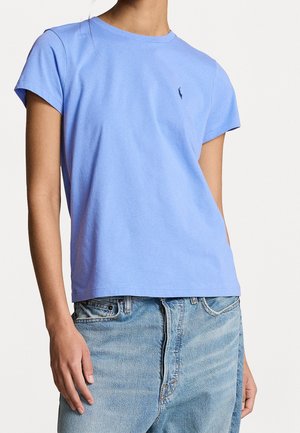 Persoon draagt een lichtblauw T-shirt met korte mouwen en een klein donker logo op de borst, gecombineerd met lichtblauwe spijkerbroek.
