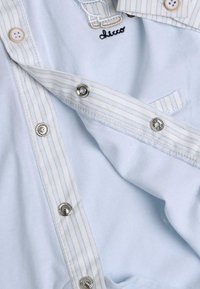 Indumento azzurro chiaro con colletto a righe bianche e bottoni a pressione argentati. Presenta un logo ricamato e una texture in tessuto morbido.