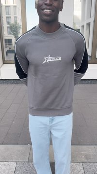 Grau sweatshirt mit schwarzen Ärmeln und einem weißen Logo, kombiniert mit hellblauen Jeans. Weiche Textur, lockere Passform und Rundhalsausschnitt.