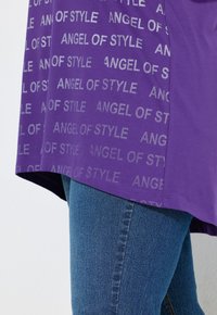 T-shirt viola con la scritta “ANGEL OF STYLE” in argento, abbinato a jeans blu. La maglietta ha una vestibilità comoda e un orlo curvo.