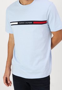 Camiseta de algodón azul claro con un diseño de rayas horizontales en negro, blanco y rojo, con el logo "TOMMY HILFIGER" centrado en el pecho.