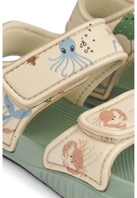 Heledad beežid sandaalid ookeaniteemaliste mustritega, millel on sinised kaheksajalgsed ja roosad krabid, reguleeritavad rihmad ning rohelised tekstureeritud tallad.