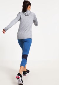 Felpa atletica grigia a maniche lunghe con cappuccio, abbinata a leggings blu con texture caratterizzati da un pannello blu scuro e scarpe da corsa nere.