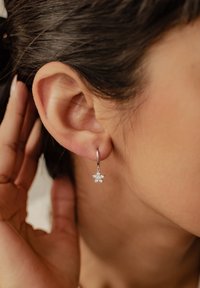 Boucle d'oreille en argent avec un charme en forme d'étoile orné de plusieurs pierres précieuses claires, suspendue près de l'oreille d'une personne. Texture lisse.