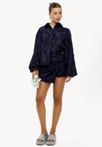 Blouse bleu marine texturée avec des manches bouffantes, associée à une jupe assortie. La tenue est complétée par une pochette bleu clair et des sandales grises à enfiler.