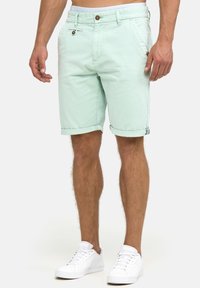 Mintgröna bomullsshorts med knappstängning, framfickor och upprullade fållar, kombinerade med vita sneakers. Slät textur och avslappnad design.