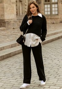 Pull noir court sur une chemise blanche, pantalon large noir et baskets blanches. Petit sac noir avec une bandoulière en chaîne. Fond en pavés.