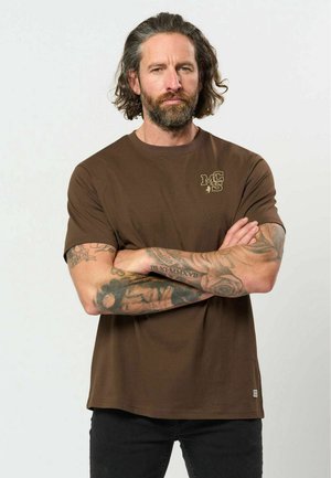 Homme aux cheveux bouclés mi-longs et barbe, portant un t-shirt marron et un pantalon noir, bras croisés montrant des tatouages couvrant toute la manche.